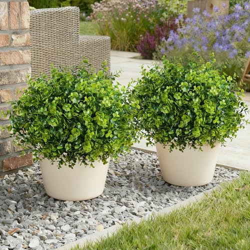 Two artificial boxwood topiary bushes in round white pots are displayed outdoors on a gravel surface, next to a wicker chair.