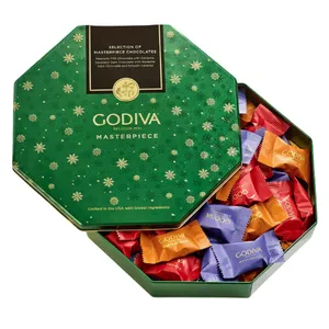 Godiva Masterpiece chocolates in a festive green hexagonal tin featuring individually wrapped pieces in red, orange, and purple. Contains milk chocolate with ganache, dark chocolate with caramel, and dark chocolate with smooth caramel.