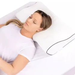 A woman is lying on her back on a bed, using a contoured white pillow with black piping for support. She wears a white t-shirt and appears relaxed.