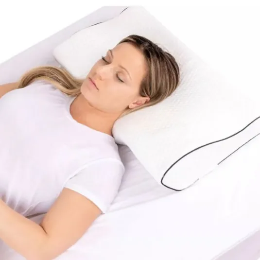A woman is lying on her back on a bed, using a contoured white pillow with black piping for support. She wears a white t-shirt and appears relaxed.