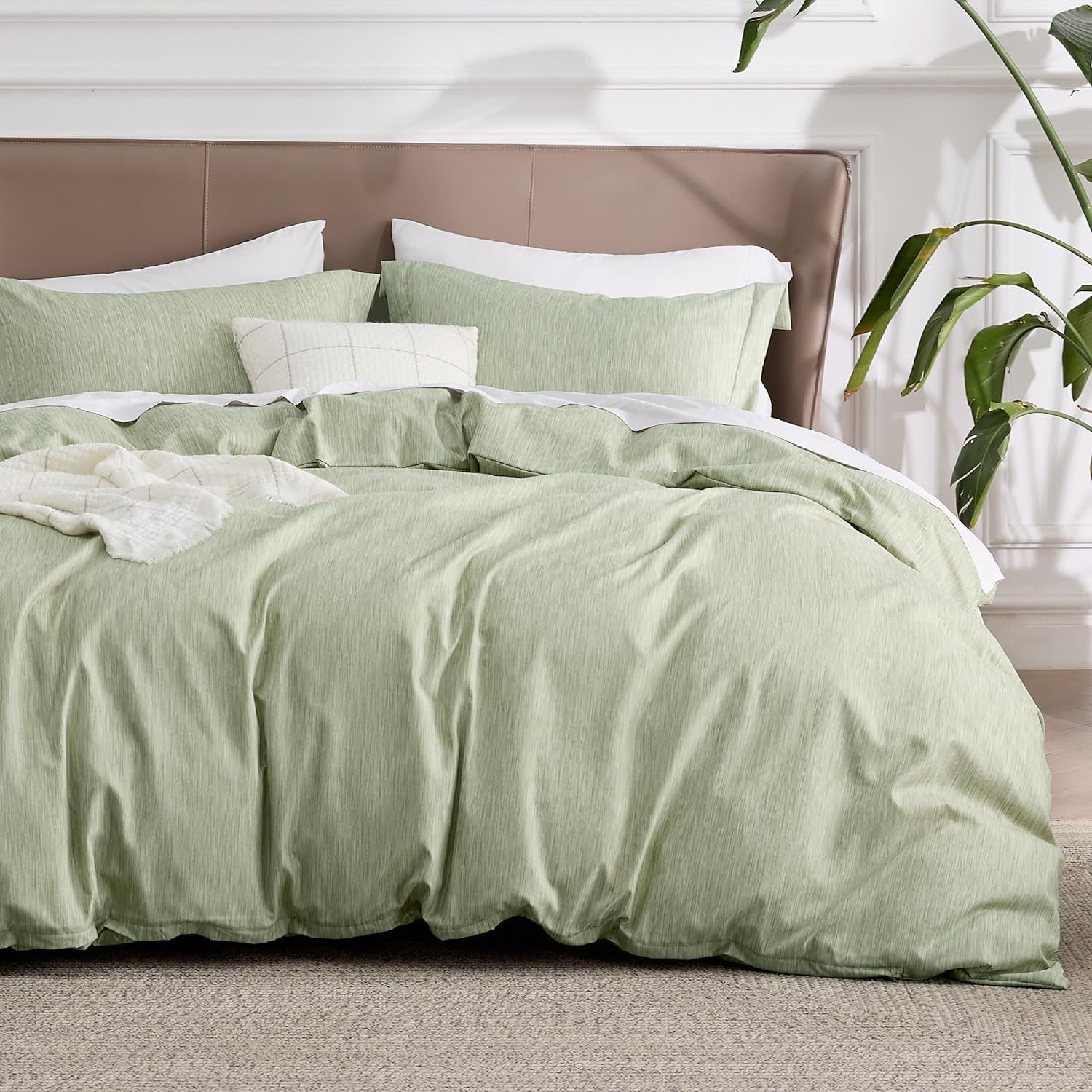 A neatly made bed features a set of light green bedding, including a duvet cover and pillowcases, with a beige headboard and a decorative white cushion. A plant is visible on the right, adding a touch of greenery to the room.
