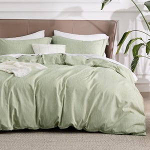 A neatly made bed features a set of light green bedding, including a duvet cover and pillowcases, with a beige headboard and a decorative white cushion. A plant is visible on the right, adding a touch of greenery to the room.