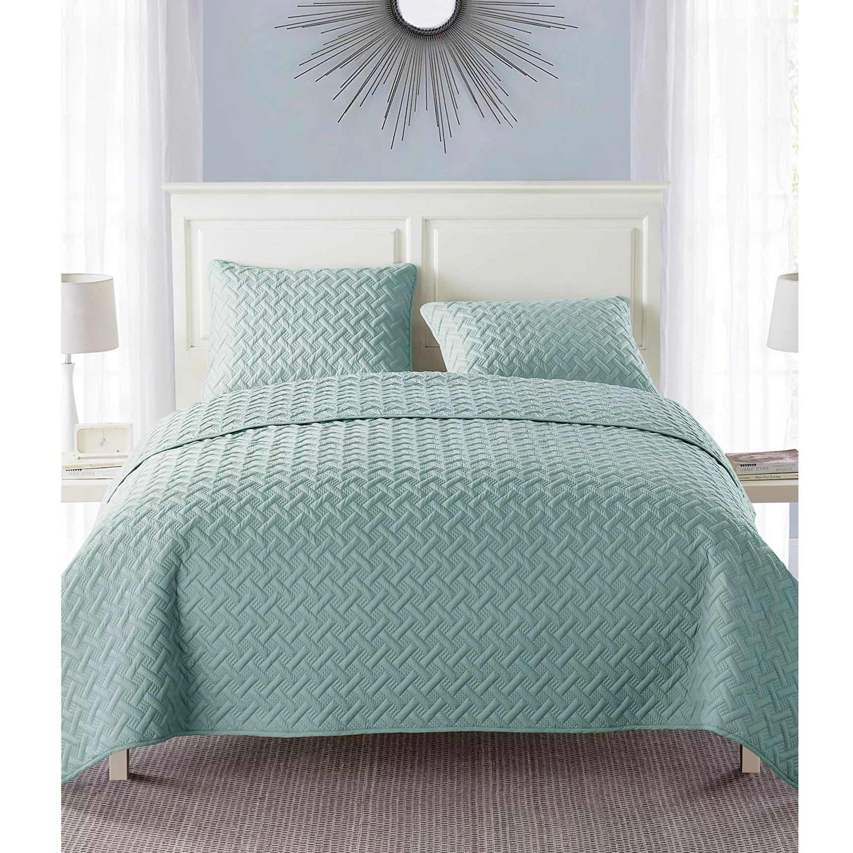 A bedding set featuring a light teal quilt with a geometric pattern and matching pillow shams on a bed. Includes a white headboard, two bedside lamps, and a decorative wall mirror.