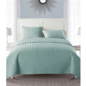 A bedding set featuring a light teal quilt with a geometric pattern and matching pillow shams on a bed. Includes a white headboard, two bedside lamps, and a decorative wall mirror.