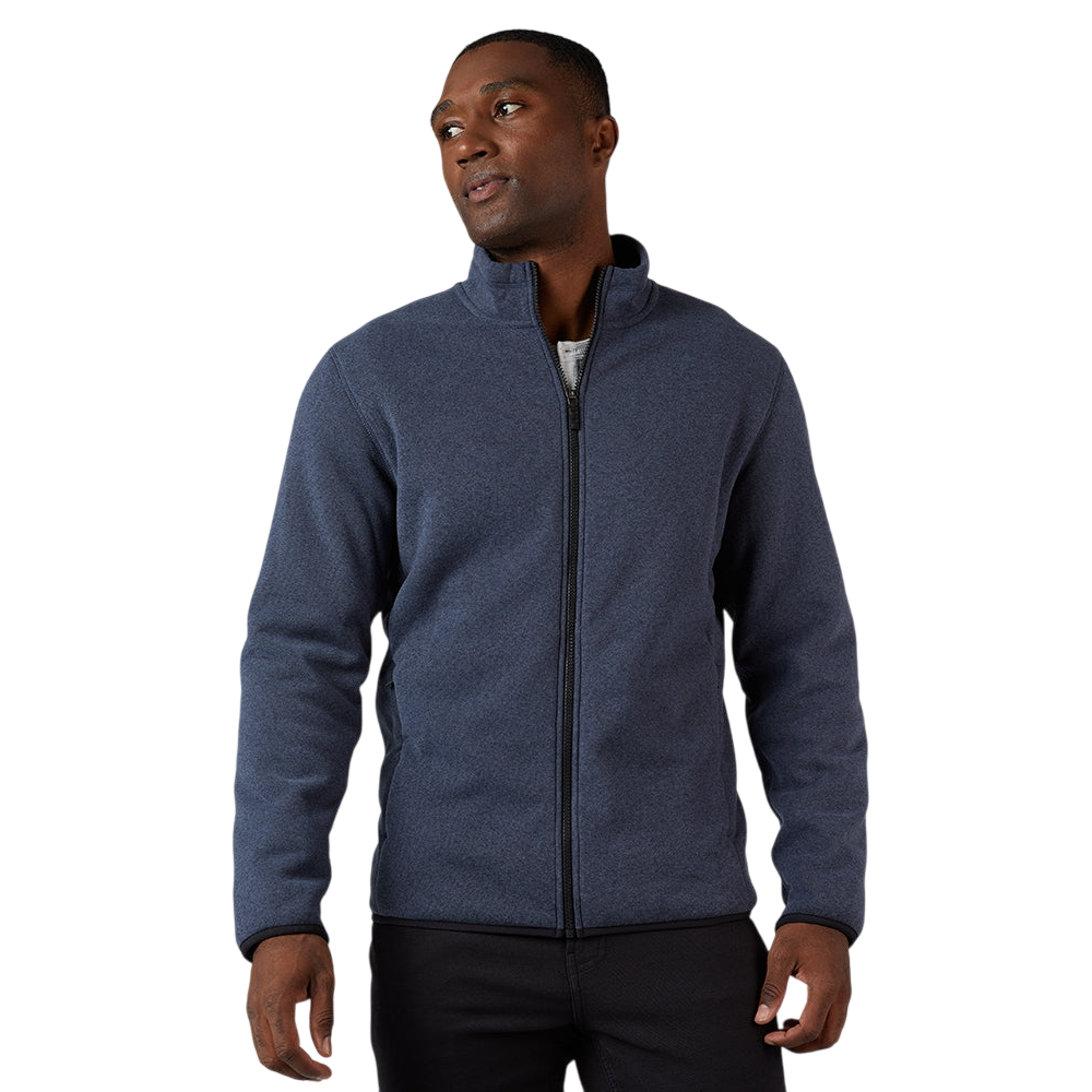 A man is wearing a navy blue zip-up jacket with a high collar, casually styled over a white shirt and black pants.