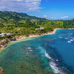 A scenic coastal view featuring a resort with palm-lined beaches, turquoise waters, and lush green hills in the background.