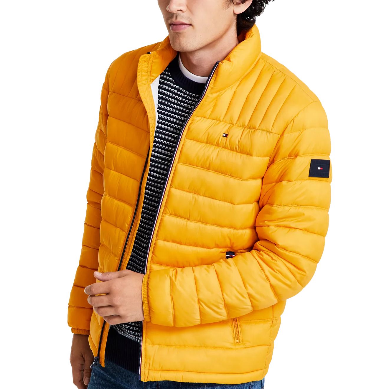 A man is wearing a bright yellow quilted jacket with small logo patches on the chest and sleeve. Underneath, he has on a black and white patterned sweater.