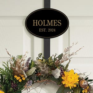 Door sign with \“HOLMES EST. 2024\“ in gold lettering is displayed above a decorative wreath featuring yellow flowers and greenery.