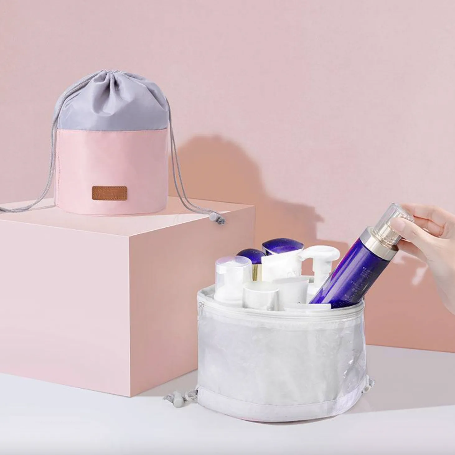 Two cylindrical drawstring cosmetic bags are displayed, one colored pink and gray, the other white, with beauty products partially inside the open white bag.
