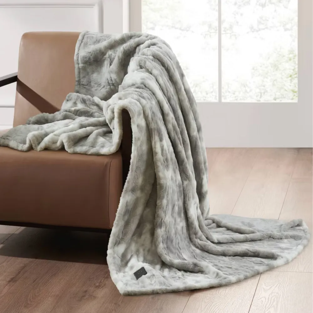 A gray faux fur throw blanket is draped over a brown leather chair with a wooden floor and large window in the background.