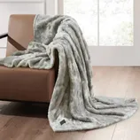 A gray faux fur throw blanket is draped over a brown leather chair with a wooden floor and large window in the background.