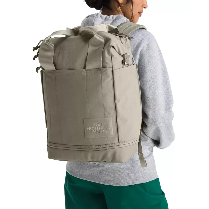 A person wearing a gray hoodie and green pants carries a beige North Face backpack featuring a roll-top design and a front logo patch.