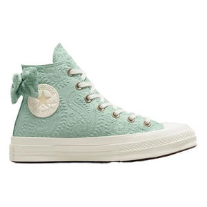 A high-top sneaker in a light pastel green with an embossed floral pattern and an attached bow near the top. It features white laces, a white rubber toe cap, and a thick white sole.