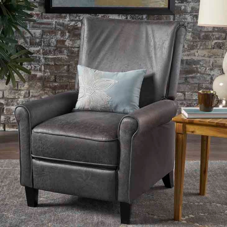 A sleek gray upholstered recliner with a plush, high back and rounded armrests, featuring a light blue pillow with an embroidered floral design. It's positioned on a rug next to a wooden side table with books and a mug.