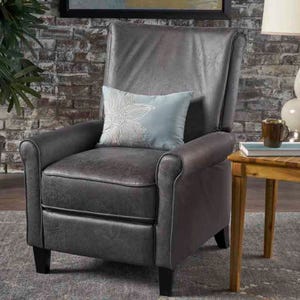 A sleek gray upholstered recliner with a plush, high back and rounded armrests, featuring a light blue pillow with an embroidered floral design. It's positioned on a rug next to a wooden side table with books and a mug.