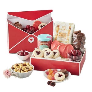 The Harry & David Valentine’s Day Box features heart-shaped cookies, chocolate-covered cherries, a jar of candy, gourmet caramel popcorn, a small bowl of colorful gummies, and cookies, all packaged in a red box designed like an envelope.