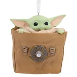 A Grogu figure ornament from \“The Mandalorian,\“ featuring the character peeking out of a brown bag with a metallic emblem.