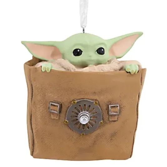 A Grogu figure ornament from \“The Mandalorian,\“ featuring the character peeking out of a brown bag with a metallic emblem.
