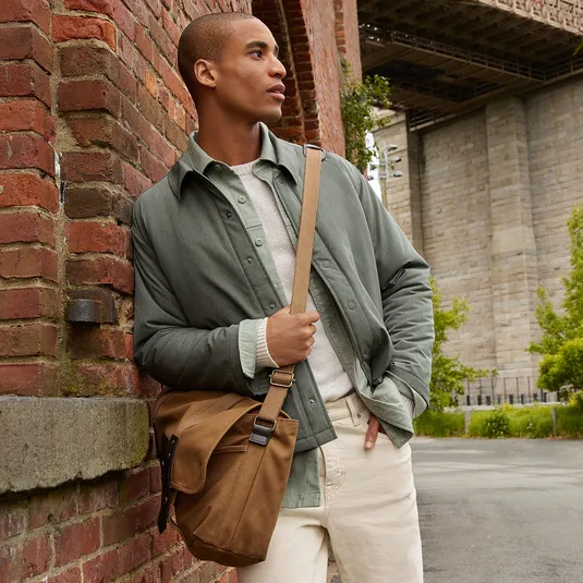 A person leans against a brick wall wearing a green jacket, layered over a light sweater and light pants, carrying a brown messenger bag.