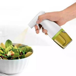 A hand is shown using a spray bottle with a transparent base, revealing a golden liquid, likely oil, being sprayed onto a salad in a white bowl.