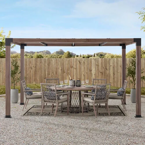 Outdoor dining set with a rectangular pergola, featuring a round table, six chairs with patterned cushions, under a partially open beam structure, set on a patterned rug, surrounded by gravel and planters.