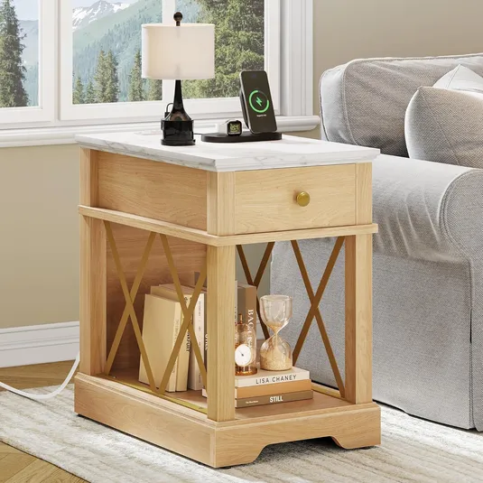 A wooden side table with a drawer and bookshelves, topped with a table lamp and phone with a charging stand. An hourglass and books are on the lower shelf.