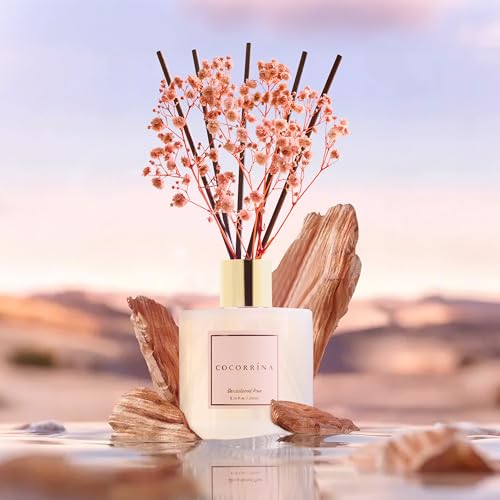 A scented diffuser with pink flowers and reed sticks in a white container labeled \“Cocorrína,\“ set against a blurred desert landscape background with stone accents.
