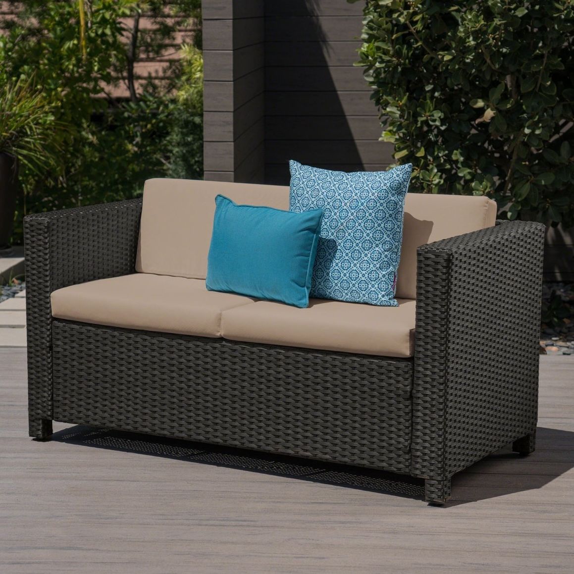 A two-seater outdoor sofa with dark wicker frame and beige cushions, featuring two blue accent pillows, one solid and one patterned.