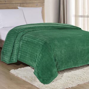 Green textured blanket draped over a bed with white pillows, placed on a white rug, in a room with a wooden headboard and light curtains.