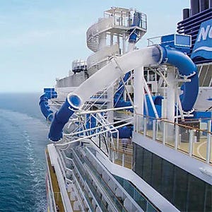 A cruise ship features a large water slide on the deck, with blue and white tubing winding around the structure. The ocean is visible in the background.