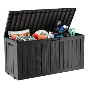 A storage box containing colorful cushions, sports balls, toys, a chainsaw, and a drill, implying versatile storage capacity. The box is black with a wood-like texture and has built-in handles and wheels.