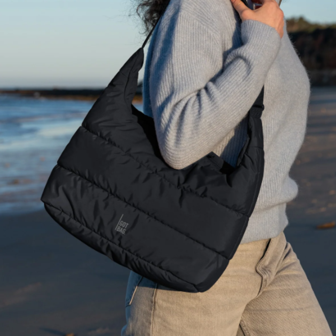 A person carries a black shoulder bag with \“GOT BAG\“ branding, wearing a gray sweater and beige pants, standing on a beach.