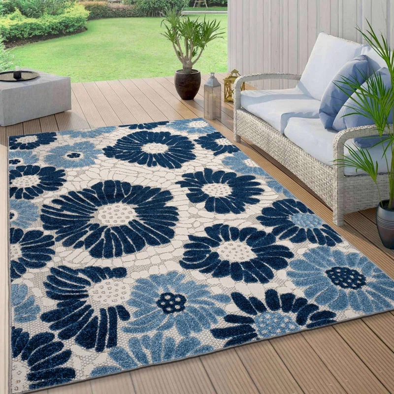 A 5' x 7' floral rug features large blue and light blue flowers on a textured beige background, suitable for both indoor and outdoor use. It complements a wicker sofa with blue cushions on a wooden patio, surrounded by potted plants.