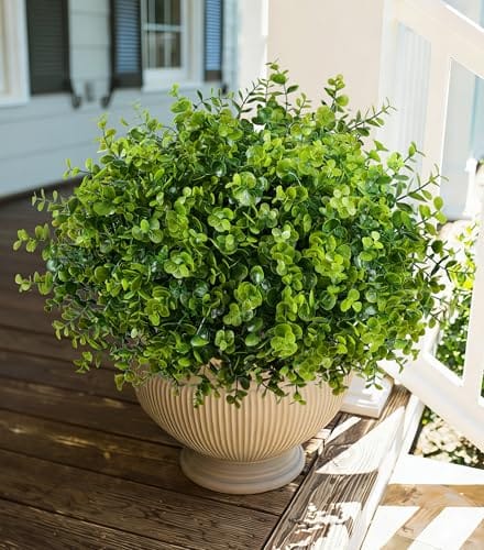 A lush, green artificial plant with small round leaves is placed in a beige, ribbed planter, sitting on a wooden deck near a white railing.