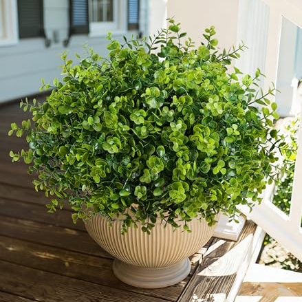 A lush, green artificial plant with small round leaves is placed in a beige, ribbed planter, sitting on a wooden deck near a white railing.
