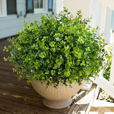 A lush, green artificial plant with small round leaves is placed in a beige, ribbed planter, sitting on a wooden deck near a white railing.