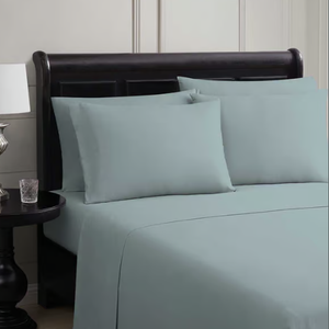 The photo shows a bed made with light blue sheets and pillowcases on a dark wooden headboard, accompanied by a bedside table holding a lamp and a small glass.