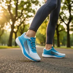 A person walking on a paved path wears blue and gray running shoes with a sleek design, surrounded by a sunlit park setting.