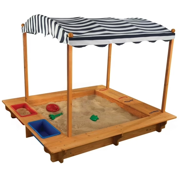 KidKraft Outdoor Covered Wooden Sandbox features a wooden frame with built-in bench seats and two plastic bins for storage, covered by a striped canopy for shade. The sandbox is filled with sand and includes a few colorful sand toys.