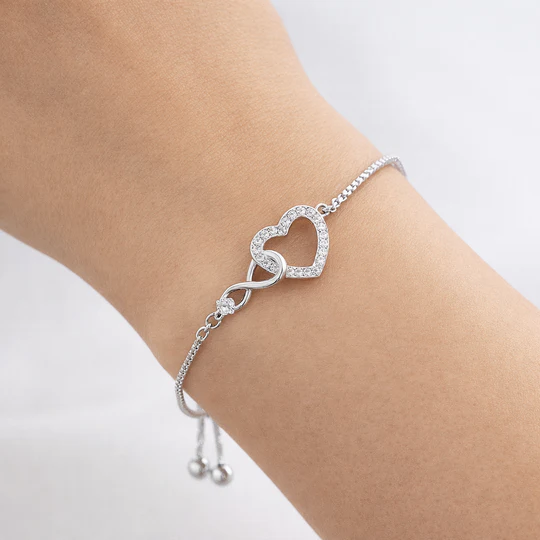 A delicate silver bracelet features an infinity symbol linked to a heart shape embellished with small clear stones, designed with an adjustable chain for a secure fit.