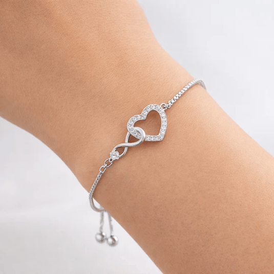A delicate silver bracelet features an infinity symbol linked to a heart shape embellished with small clear stones, designed with an adjustable chain for a secure fit.