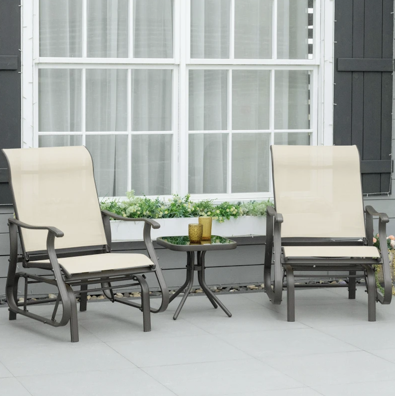 Two beige outdoor glider chairs with armrests are positioned around a small round black glass table, set on a gray patio in front of a window with a flower box.