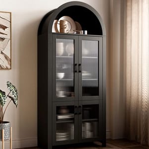 A tall, dark arched display cabinet with four glass-paneled doors and visible internal shelves, showcasing neatly arranged dishes and decor. The cabinet has a modern design and is placed against a light beige wall with a potted plant nearby.