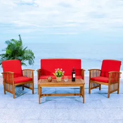 Outdoor furniture set with two red-cushioned armchairs and a loveseat, complemented by a wooden coffee table featuring a plant, glasses, and a wine bottle. The setup is by the ocean with a backdrop of foliage.
