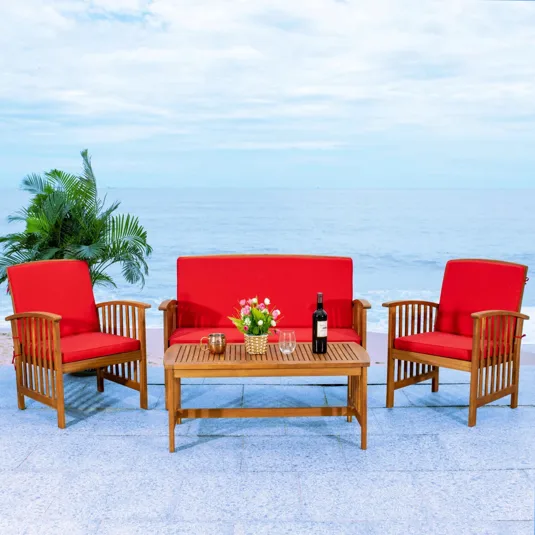 Outdoor furniture set with two red-cushioned armchairs and a loveseat, complemented by a wooden coffee table featuring a plant, glasses, and a wine bottle. The setup is by the ocean with a backdrop of foliage.