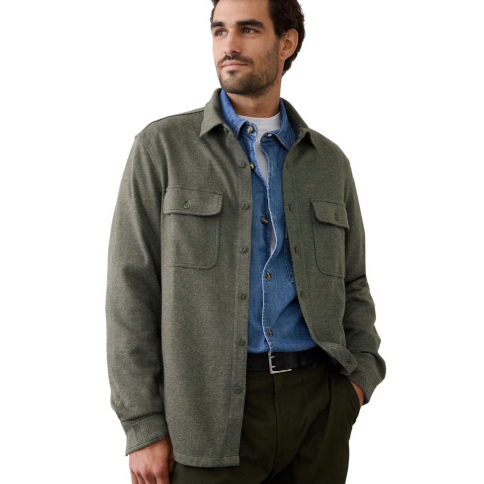 A man is wearing an olive green overshirt with buttoned pockets over a denim shirt and white tee. His outfit is paired with dark green pants and a black belt.