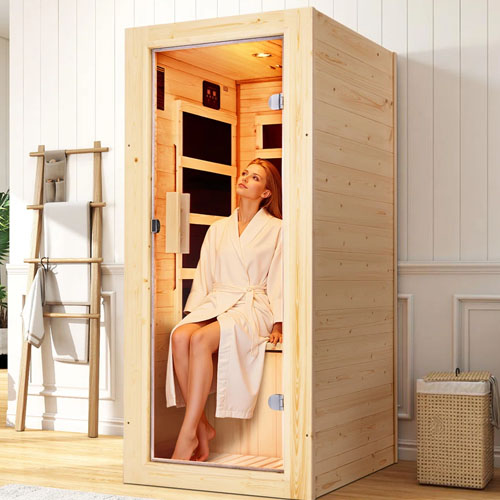 A small wooden infrared sauna with a glass door, featuring a person inside sitting on a bench, dressed in a white robe. The setup is placed in a minimalist room with a towel rack and a woven basket nearby.