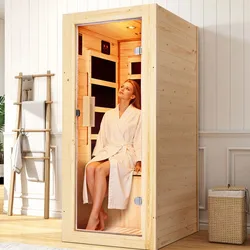 A small wooden infrared sauna with a glass door, featuring a person inside sitting on a bench, dressed in a white robe. The setup is placed in a minimalist room with a towel rack and a woven basket nearby.