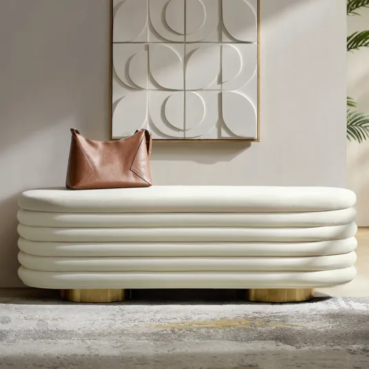 A brown leather handbag sits on a white, layered bench with gold legs, placed under a geometric white wall art piece alongside a leafy plant.