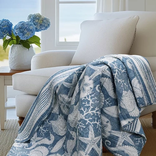 A blue and white coastal-themed quilt with starfish, seashells, and coral patterns draped over a beige armchair. Next to it is a white vase with blue hydrangeas on a side table.